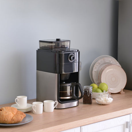 Table with a hotel breakfast scene showing a coffee machine, some mugs, croissant, apples in a bowl and plates.